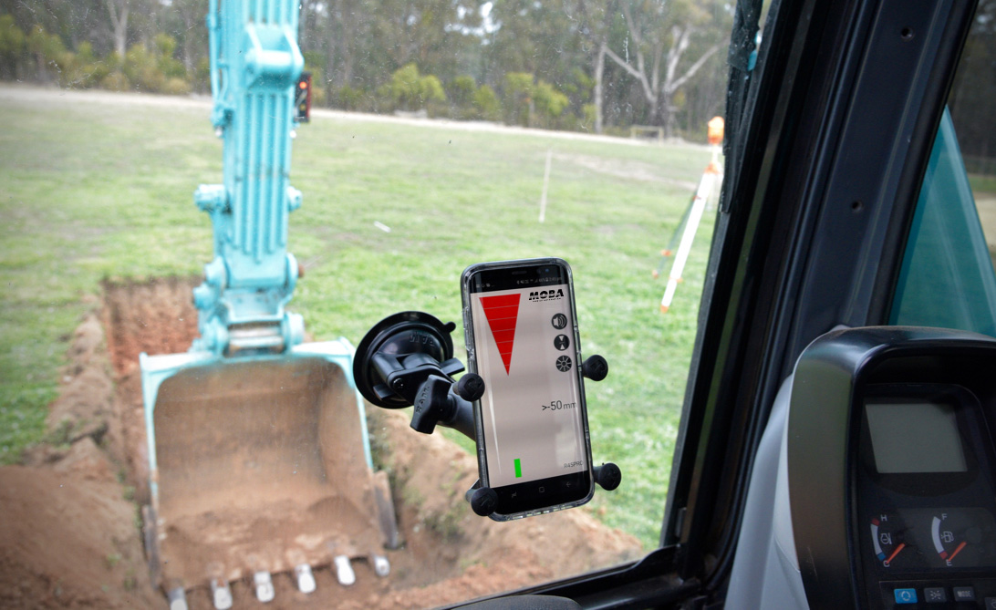 Smartphone mounted to the cab window showing a construction app, with an excavator digging outside in the background.