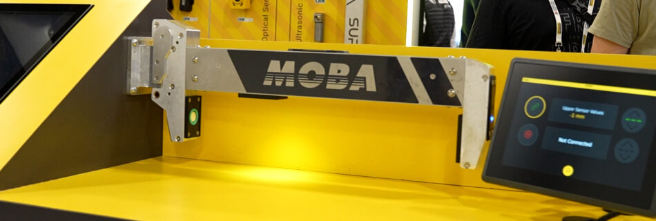 Close-up of a yellow MOBA industrial machine at a trade show, showing mounting hardware and a nearby touchscreen control panel on the right