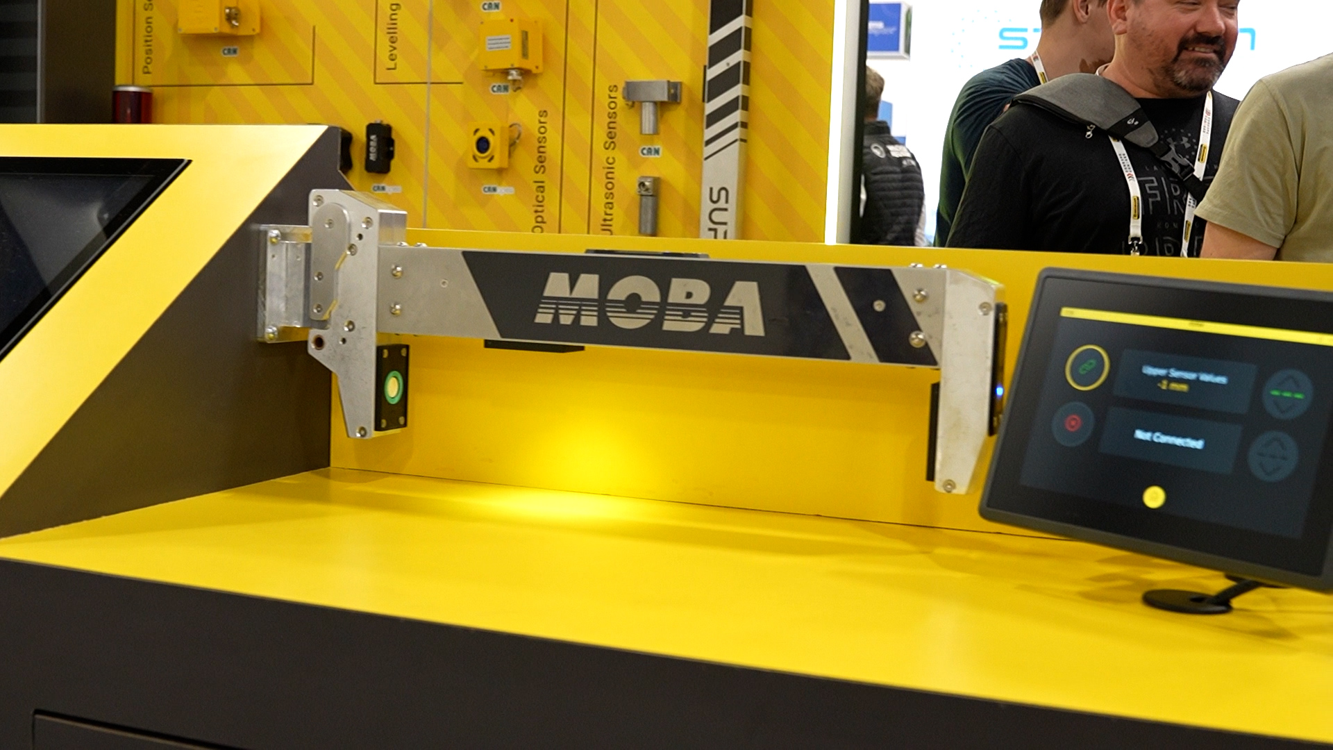 Close-up of a yellow MOBA industrial machine at a trade show, showing mounting hardware and a nearby touchscreen control panel on the right