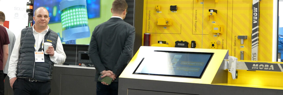 Two men stand at a trade show booth with a yellow MOBA display of sensors and a large screen showing a robotic component behind them
