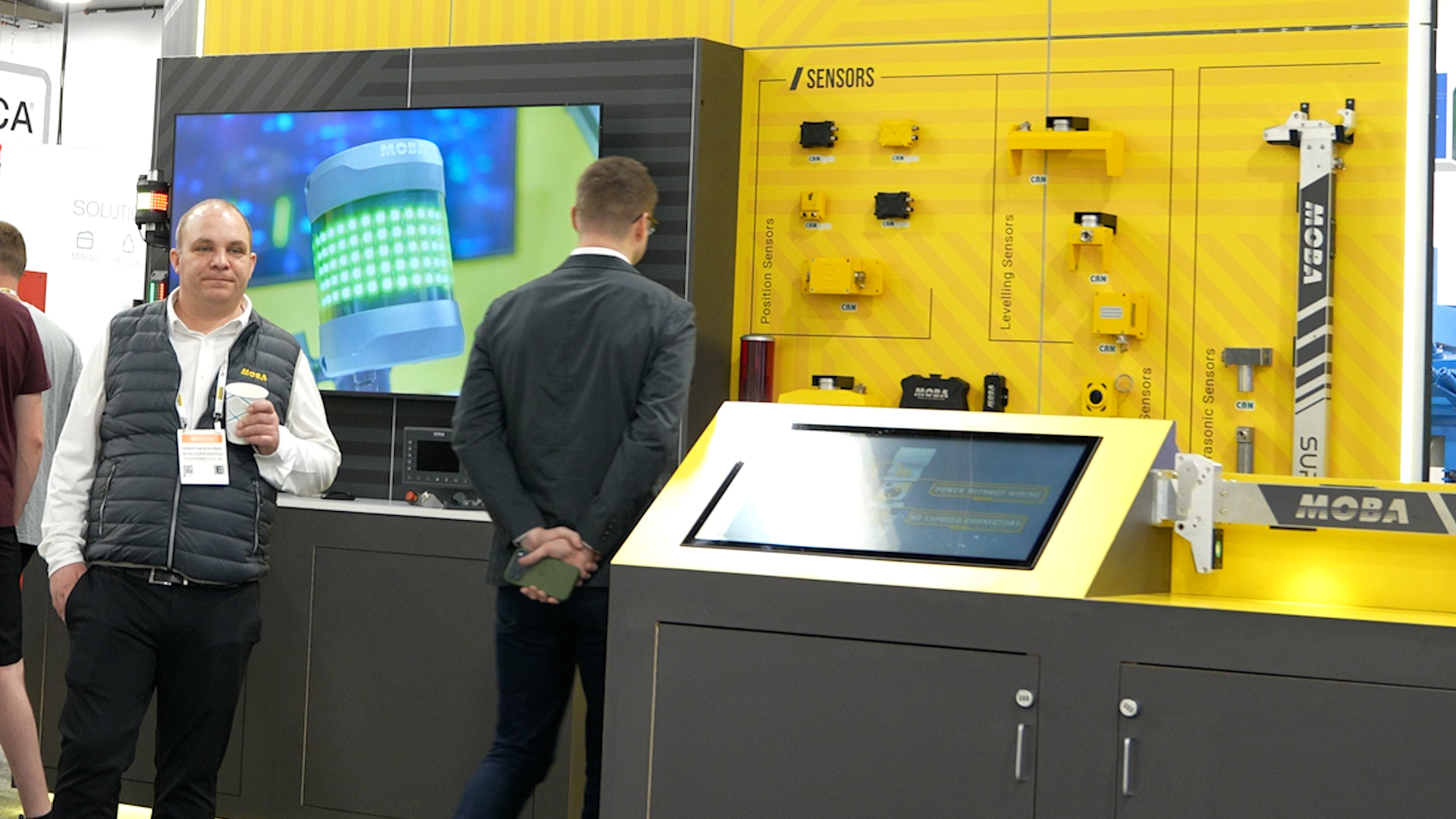 Two men stand at a trade show booth with a yellow MOBA display of sensors and a large screen showing a robotic component behind them