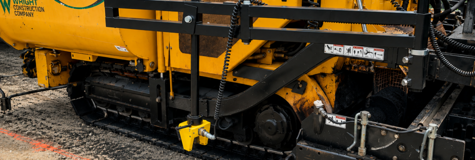 Close-up of a yellow asphalt paver with the WEILER logo and guard rails, on a construction site.