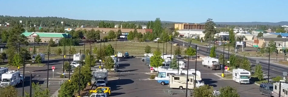 Broad view of an RV parking lot with many motorhomes and campers lined up among trees under a clear blue sky, with a commercial area in the distance.