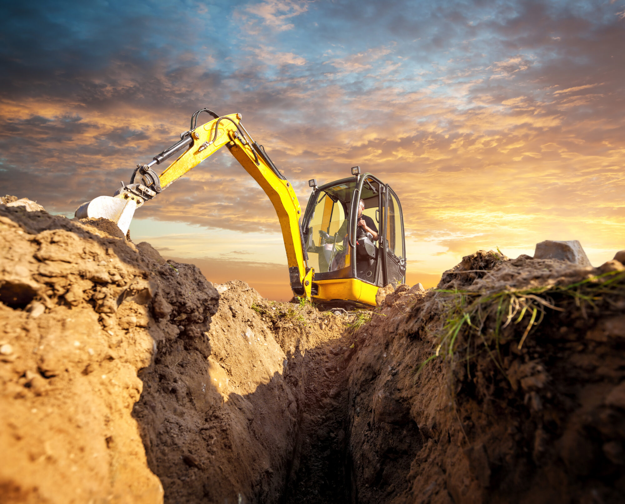 Yellow excavator with extended arm working beside a deep dirt trench at a construction site during a orange-pink sunset sky.