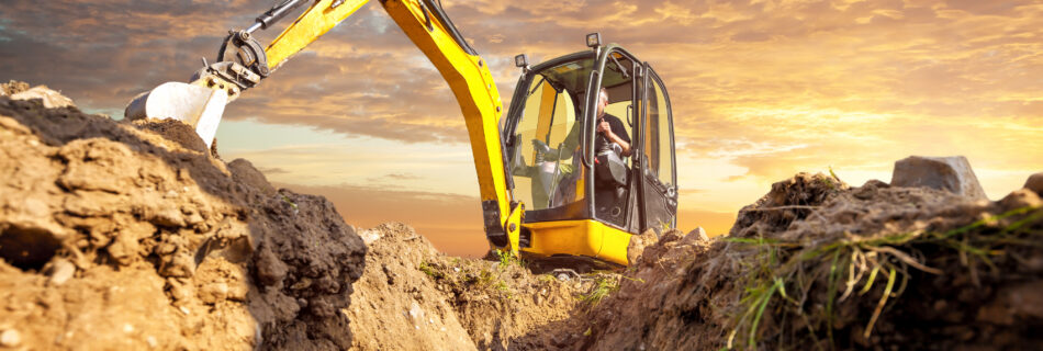 Yellow excavator with extended arm working beside a deep dirt trench at a construction site during a orange-pink sunset sky.