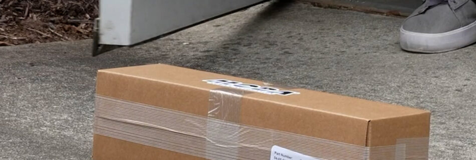 Brown cardboard shipping box sealed with tape rests on concrete ground near a gray door, with a gray sneaker visible in the corner.