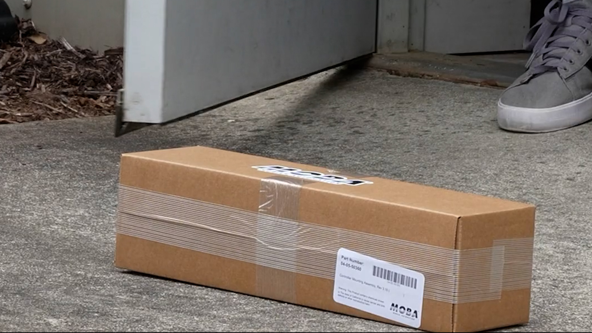 Brown cardboard shipping box sealed with tape rests on concrete ground near a gray door, with a gray sneaker visible in the corner.